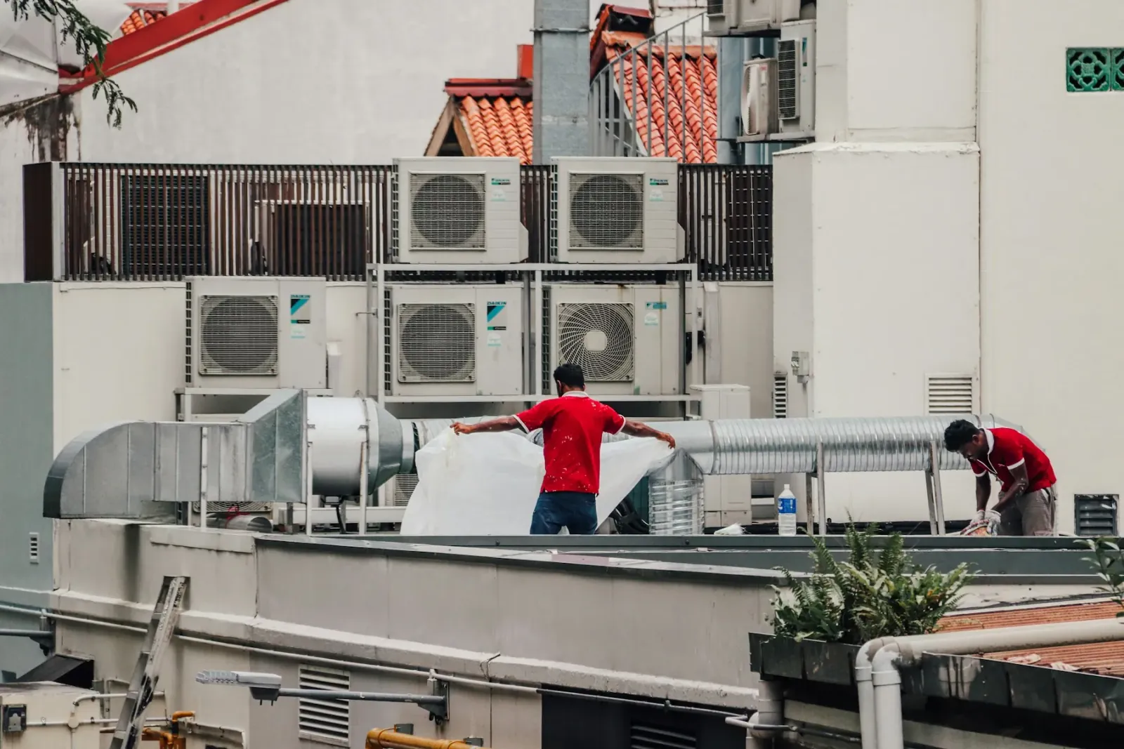 Rows of air-conditioning condenser units and spiral ductwork on a rooftop, with two workers in red shirts